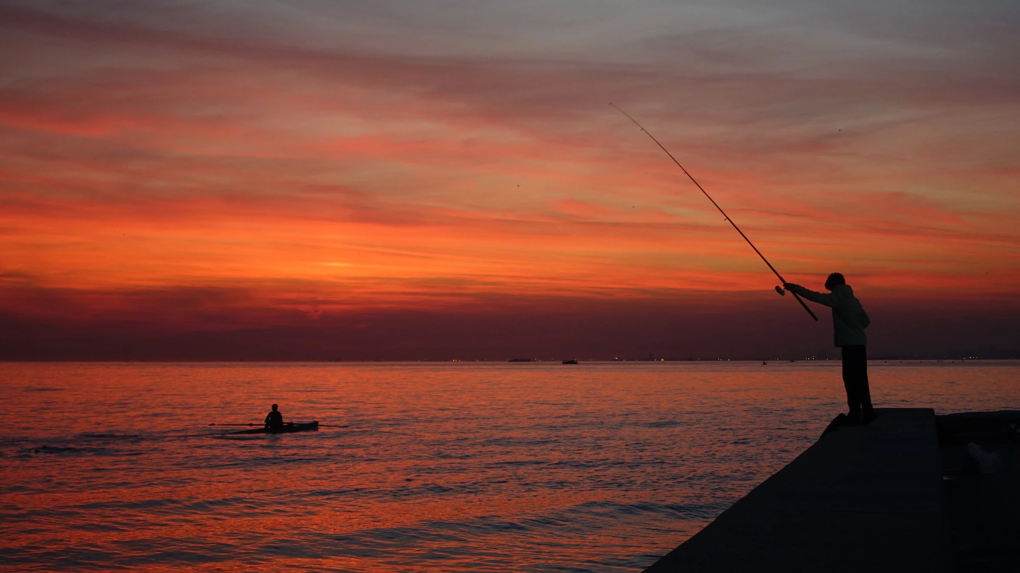 A person casts a fishing rod from the shore while a rower passes across the orange and pink sea at sunset