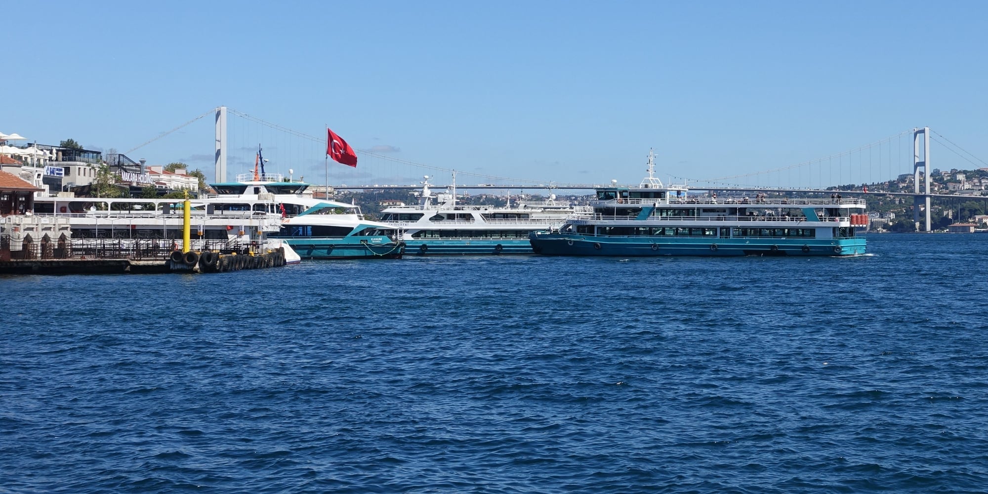 Ferries beside a waterfront pier, with blue water and a suspension bridge behind them.