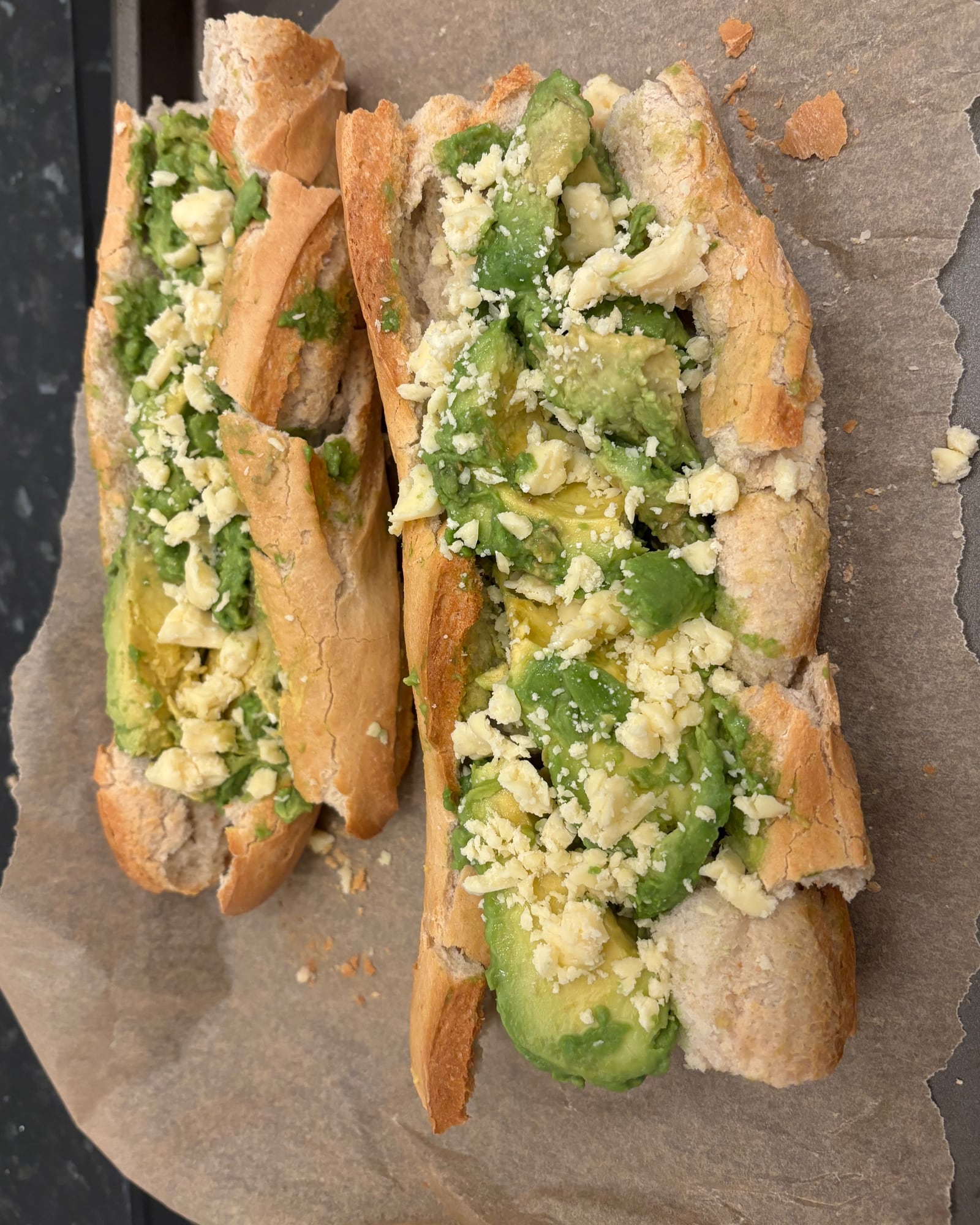 Two open baguettes filled with avocado and crumbled cheese on baking paper.