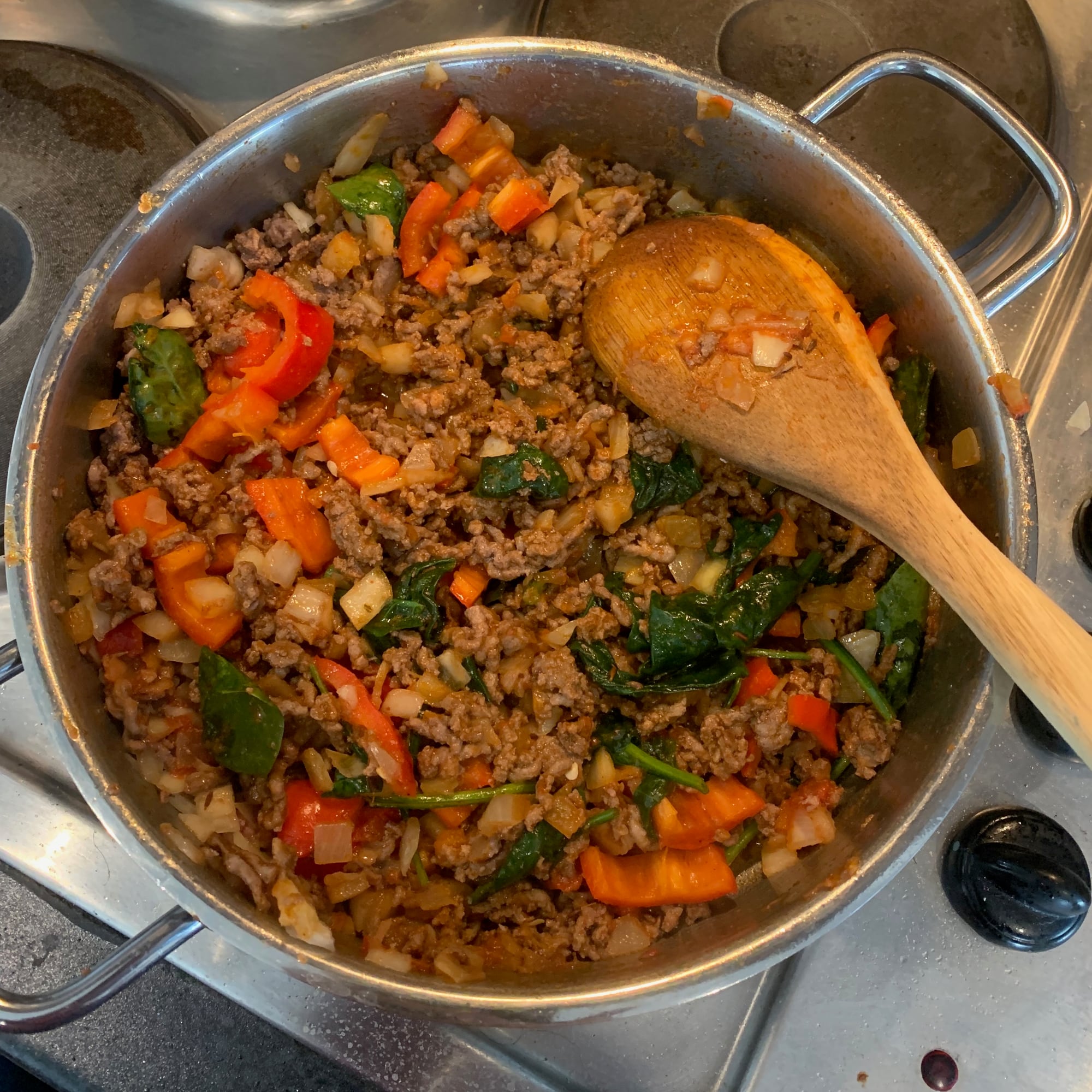 A pan of minced meat, peppers, onions, and leafy greens, with a wooden spoon.