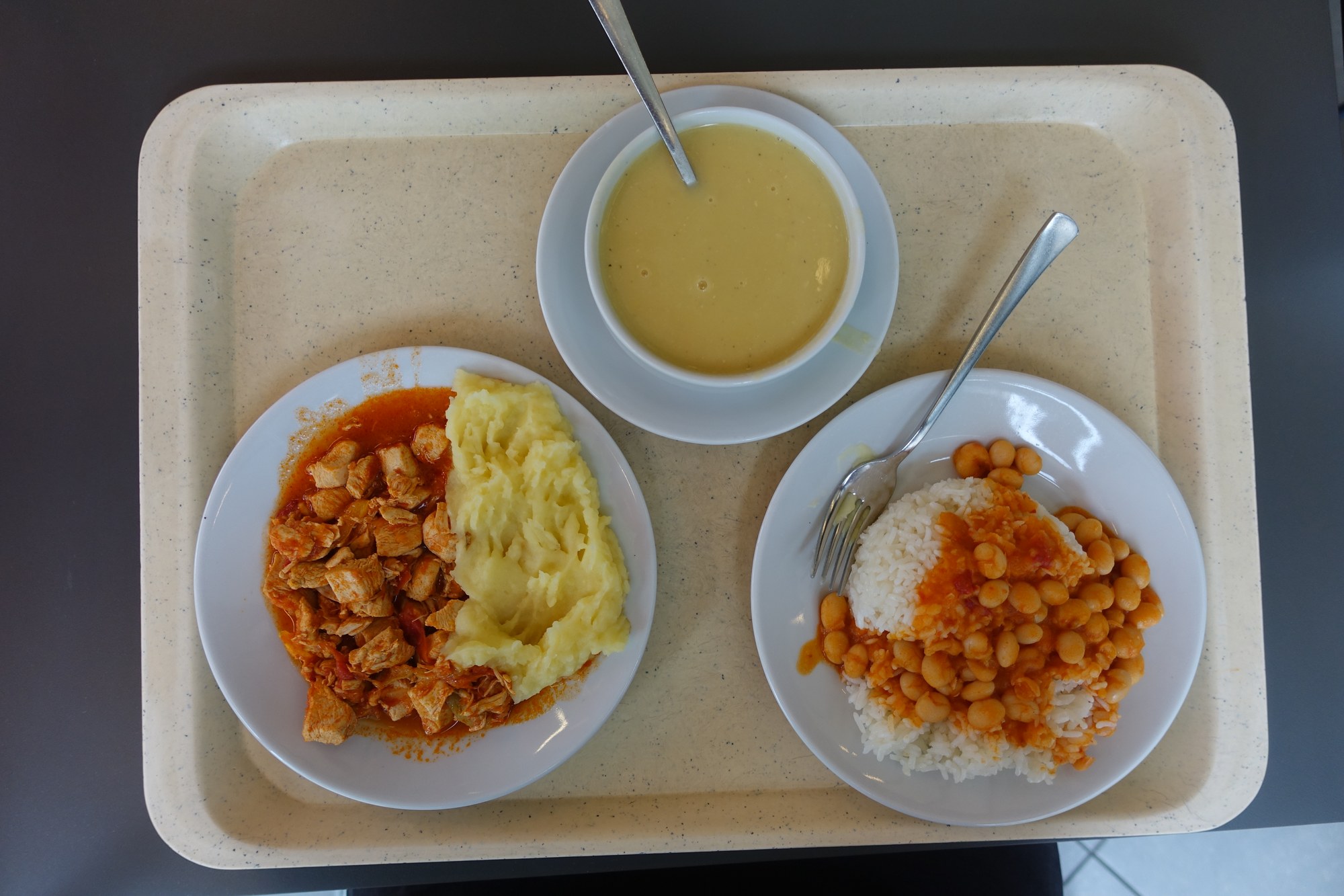 A tray with rice topped with white beans, chicken in sauce with mashed potatoes, and lentil soup.