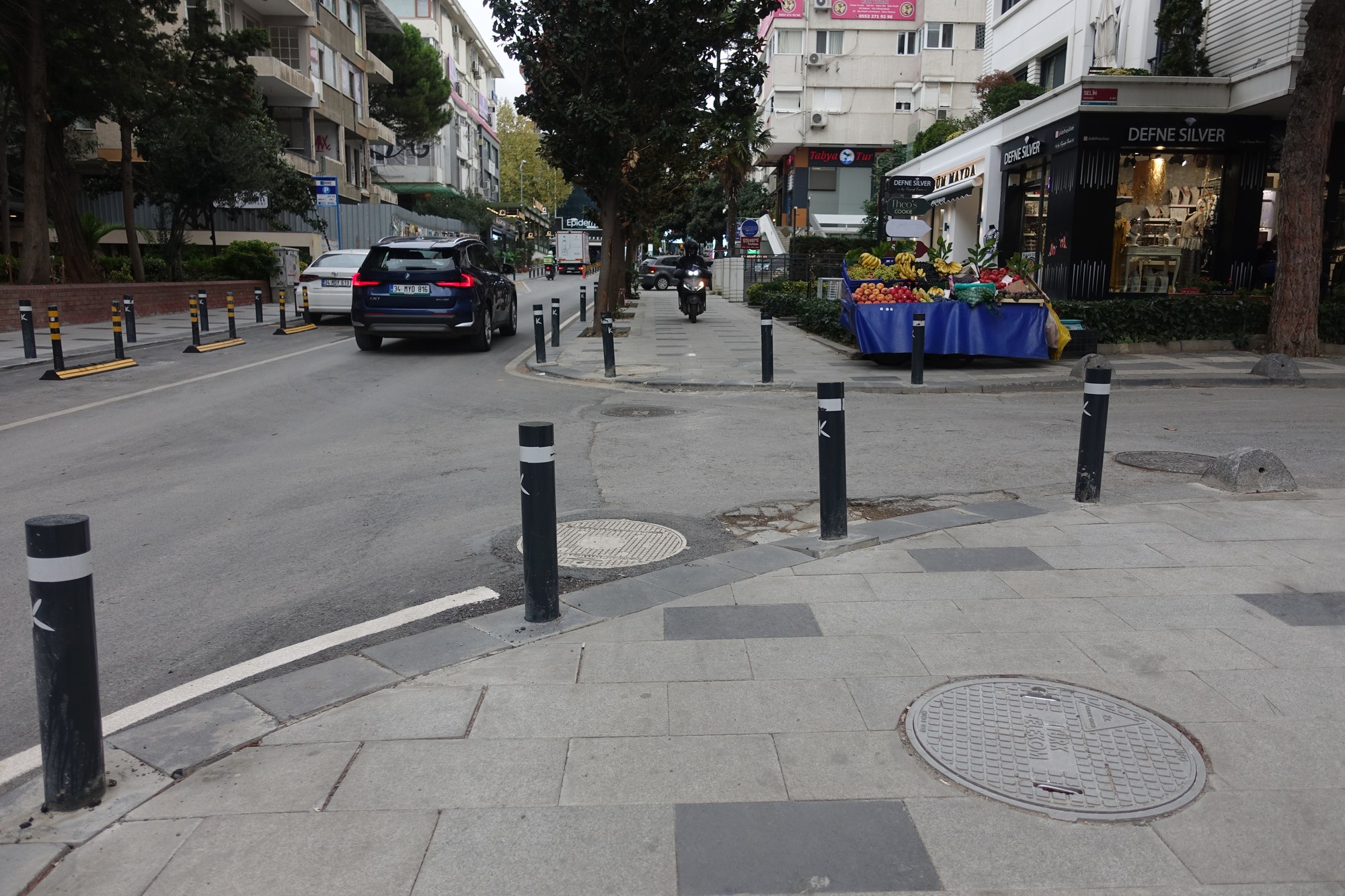 A motorcycle travels along the pavement beside a fruit stall, with bollards lining the pavement.