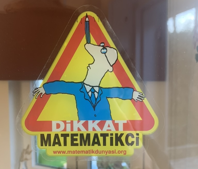 A colourful warning sign showing a mathematician holding a pen and the words Caution: Mathematician.